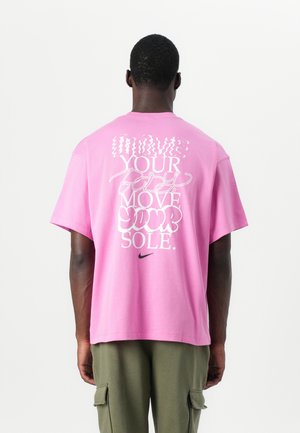 Mężczyzna w różowej koszulce Nike z białym napisem „MOVE YOUR body MOVE YOUR SOLE.” i oliwkowych spodniach bojówkach, stoi tyłem do kamery.