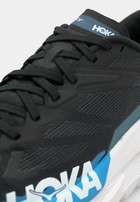 Czarne buty biegowe z teksturowaną cholewką, wyposażone w białą i niebieską podeszwę oraz logo, a także kontrastującymi czarnymi sznurówkami dla pewnego dopasowania.