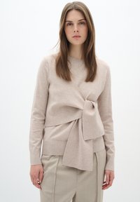 Beige gestrickter Pullover mit Knoten-Detail vorne, Rundhalsausschnitt und langen Ärmeln. Kombiniert mit hellen, taillierten Hosen.
