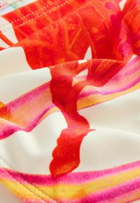 Gros plan d'un tissu avec des motifs floraux abstraits rouges, roses et jaunes sur fond blanc, montrant la texture et les détails des coutures.