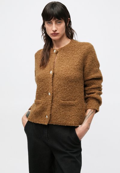Brązowy, miękki kardigan z okrągłym dekoltem, zapinany na guziki, z dwoma kieszeniami z przodu. Nosi się go z czarnymi spodniami, posiada teksturowaną powierzchnię.
