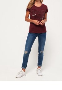 Camiseta ajustada burdeos con lettering y gráficos texturizados, combinada con jeans azules desgastados y zapatillas blancas.