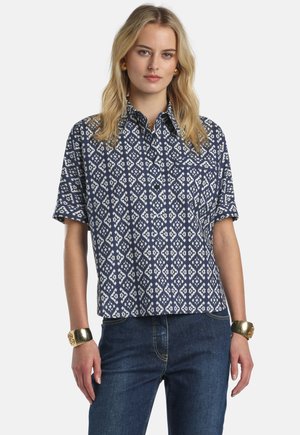 Blondinka, ki nosi temnomodro in belo srajco s kratkimi rokavi z vzorcem ter modre kavbojke, okrašena z zlatimi zapestnicami in uhančki.