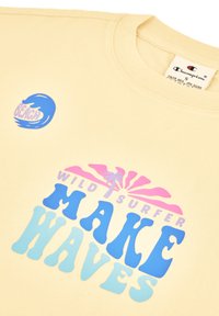 Camiseta de algodón beige con cuello redondo. Presenta texto impreso en pantalla colorido y gráficos de olas en azul y rosa. Etiqueta de tamaño visible.