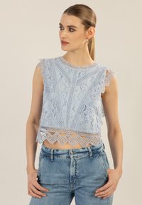 Helles blauer Spitze-Crop-Top mit gewellten Kanten, filigranen Mustern und kurzen Ärmeln, kombiniert mit hellblauen Jeans.