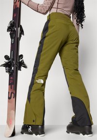Grüne Skihosen mit schwarzen Seiteneinsätzen, aus strapazierfähigem Material. Mit dem Logo von The North Face und Knopfverschluss an den Knöcheln. Skiausrüstung ist sichtbar.