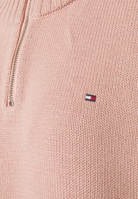 Lichtroze gebreide trui met een kwart-rits halslijn en een textuurstof. Voorzien van een klein geborduurd logo in rood, wit en blauw.