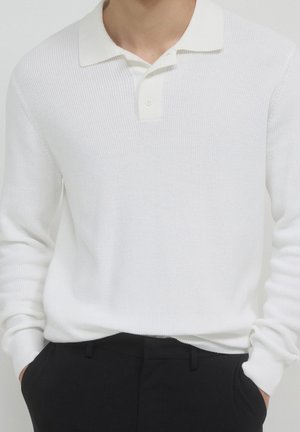 Homme portant un polo blanc en maille à manches longues rentré dans un pantalon noir, les mains dans les poches, sur un fond uni.