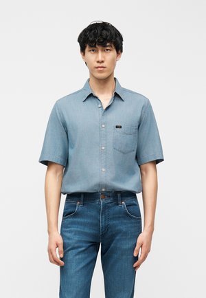 Fiatal férfi áll, rövid ujjú világoskék gombos ingben és középkék farmernadrágban, egyszínű háttér előtt.