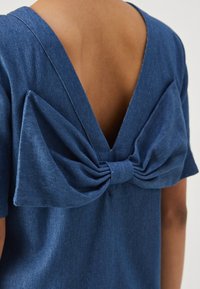 Σκούρο μπλε τζιν τοπ με βαθύ V στην πλάτη, διακοσμημένο με μια μεγάλη κορδέλα από το ίδιο ύφασμα, που δημιουργεί μια υφασμάτινη υφή.