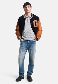 Veste universitaire marine et beige avec des manches en cuir, lettre "G", polo gris, jeans bleu délavé et chaussures noires. Se tenant avec les mains dans les poches.