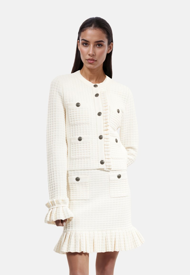 Costume en tricot de couleur crème avec un motif texturé, comprenant une veste à boutons et une jupe à volants. Comprend quatre poches et des boutons en métal.