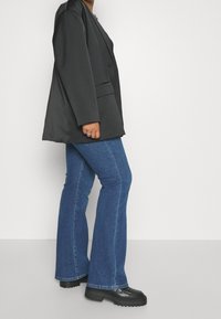 Personne portant un blazer noir surdimensionné, un jean flare bleu, des chaussures à plateforme noires et une bague à la main gauche, sur un fond blanc uni.