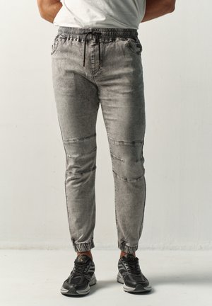 Hombre con vaqueros jogger grises con puños elásticos y zapatillas negras, de pie frente a un fondo claro y liso.