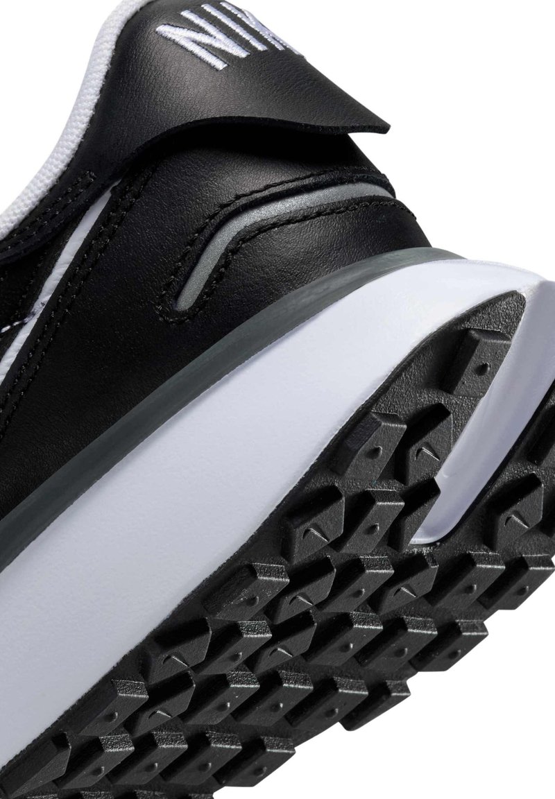 Zapatilla deportiva negra con una suela de goma texturizada con patrones triangulares, parte superior de cuero elegante y acentos blancos a lo largo de la entresuela.