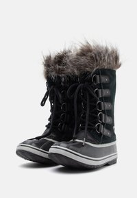 Bottes en daim noir avec bord en fausse fourrure, accents en caoutchouc et lacets à l'avant. Semelle texturée pour une meilleure adhérence, avec des rayures grises sur le dessous.