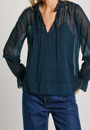 Vrouw draagt een doorschijnende marineblauwe blouse met getextureerde stippen, een ruches kraag, striklinten en lange pofmouwen, gecombineerd met donkerblauwe spijkerbroek.