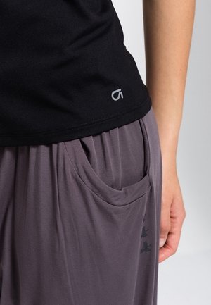 Nahaufnahme einer Person, die ein schwarzes Hemd und lockere graue Hosen mit einer Fronttasche und einem dezenten Logo auf der Hose trägt.
