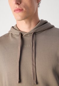 Lys brun hettegenser laget av mykt materiale, med justerbar hette og en liten "HUGO" logo i en matchende nyanse foran.
