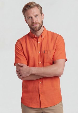 Homme barbu aux cheveux châtain clair portant une chemise orange à manches courtes boutonnée et un pantalon beige, bras croisés, regardant la caméra.
