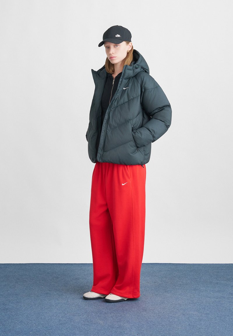 Puffer jacket in dark green, zippered front, hooded; red sweatpants with white logo. Model wears black cap and gray slip-on shoes.