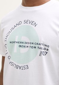 Hvid bomulds t-shirt med et rundt grafisk design i blå og mintgrønne nuancer, tekst der fremhæver brandnavnet og etableringsåret.