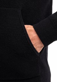 Schwarzes gestricktes Kleidungsstück mit einem strukturierten Muster. Verfügt über eine Tasche, die mit einer sichtbaren Hand ausgerichtet ist, und zeigt dicke, gerippte Bündchen sowie einen Saum.