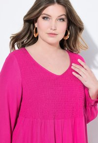 Blusa fucsia con scollatura a V, caratterizzata da una trama smock sul corpino e maniche lunghe, tessuto liscio e vestibilità rilassata.