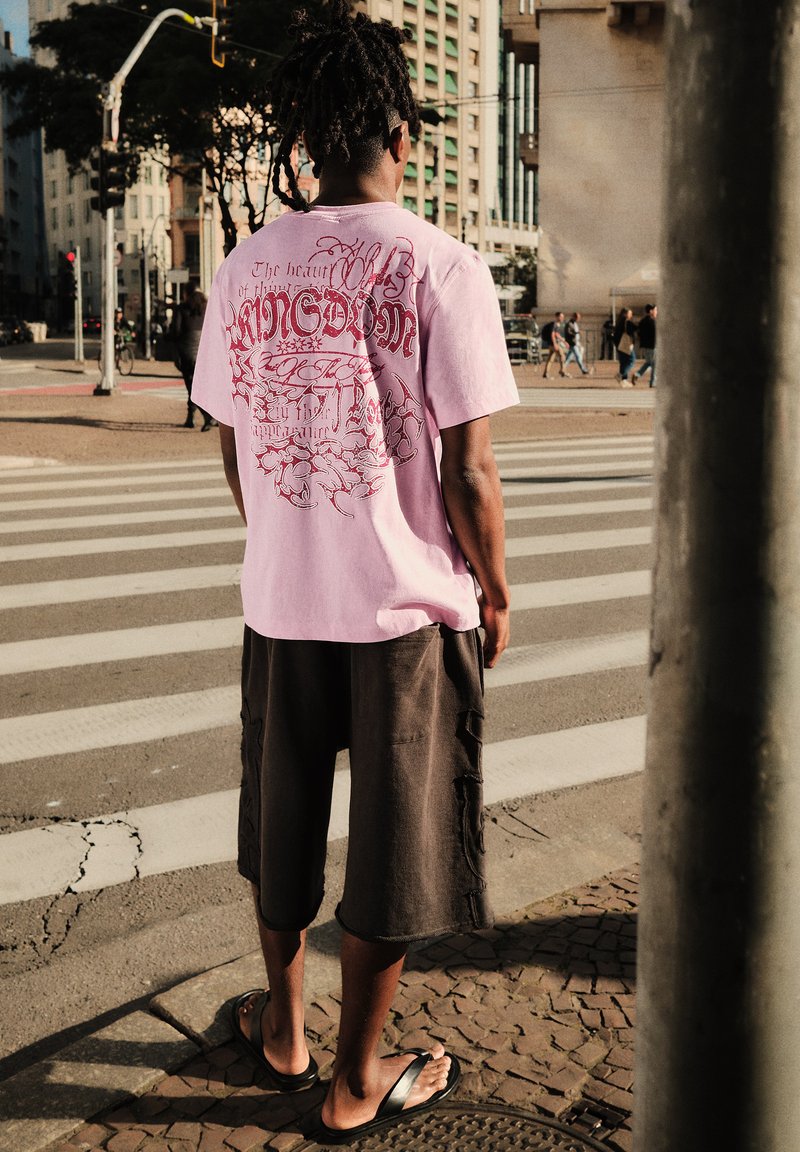 Personne avec des dreadlocks portant un t-shirt rose à motif graphique, un short foncé et des sandales, se tient à un passage piéton en zone urbaine.