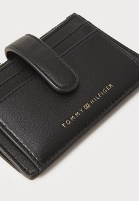 Portefeuille en cuir noir avec une surface texturée, fermé par un rabat magnétique, doté de plusieurs emplacements pour cartes et d'un accent logo doré.