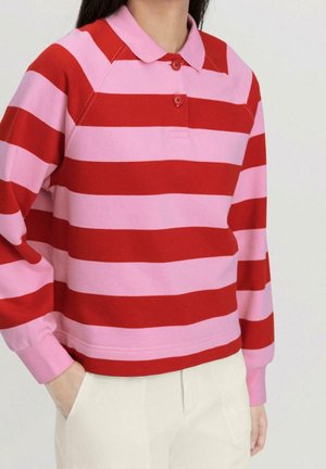 Personne portant un polo à manches longues rayé horizontalement rose et rouge avec des boutons et un pantalon blanc, une main dans la poche.