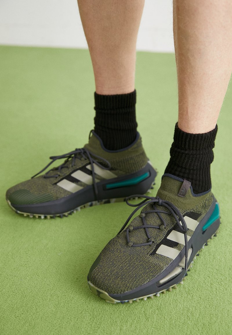 Zapatillas deportivas de punto verde con acentos grises, que cuentan con un tejido texturizado, suela de goma y tres franjas negras a un lado. Calcetines negros con canalé.