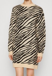 Femme portant une robe pull à manches longues imprimée zèbre beige et noire avec poignets et ourlet côtelés, debout devant un fond clair uni.