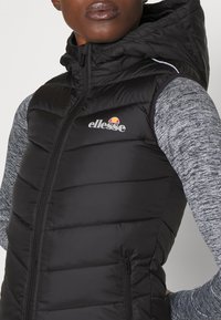 Svart vadderad ärmlös väst med huva, med en framdragkedja och logotypen "ellesse." Grå texturerade långa ärmar syns under.