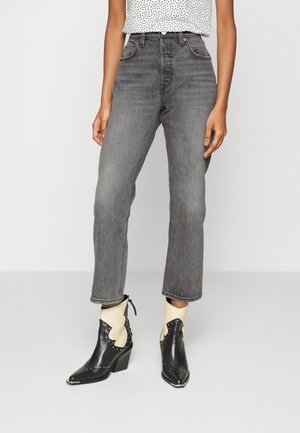 Personne portant un jean noir délavé taille haute et des bottes de cowboy pointues noires et blanches avec des clous, debout devant un fond uni.