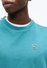 Κοντινό πλάνο του λαιμού και του άνω στέρνου ενός ατόμου που φοράει τιρκουάζ φούτερ Puma πάνω από λευκό πουκάμισο, με το λογότυπο Puma στο στήθος.