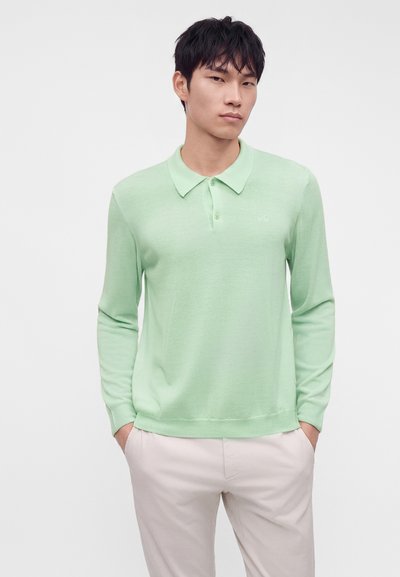 Jeune homme aux cheveux courts et foncés portant un polo manches longues vert clair et un pantalon clair, mains dans les poches, fond neutre.