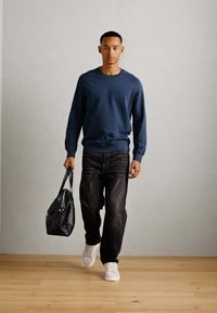 Marinegenser, mørke jeans og hvite sneakers. Bærer en svart skulderveske. Står på lyse tregulv med en enkel grå bakgrunn.