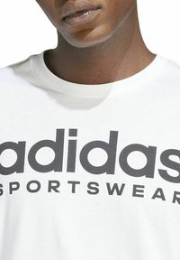 Camiseta de algodón blanca con un gran logo negro de "adidas" y el texto "SPORTSWEAR" debajo. Diseño de cuello redondo y mangas cortas.