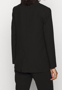 Blazer noir sur mesure avec une coupe droite, une fente simple à l'arrière, et des épaules structurées. Confectionné dans un tissu lisse.