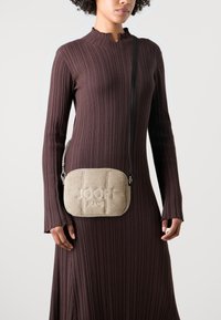 Borsa a tracolla trapuntata beige con ricamo "JOOP! JEANS". Presenta una tracolla a catena, una consistenza morbida e una forma arrotondata, portata all'anca.