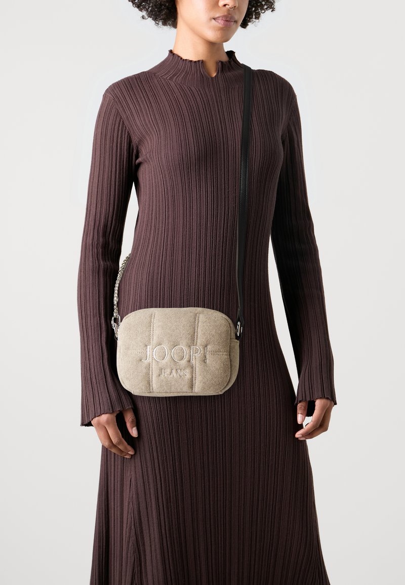 Borsa a tracolla trapuntata beige con ricamo "JOOP! JEANS". Presenta una tracolla a catena, una consistenza morbida e una forma arrotondata, portata all'anca.