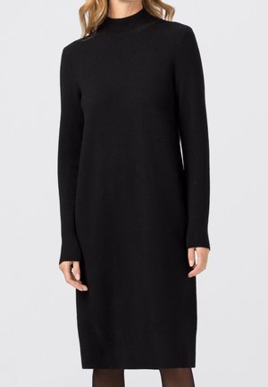 Femme portant une robe-pull noire unie à manches longues et longueur genoux, avec un col montant et des poignets côtelés, sur un fond gris clair.
