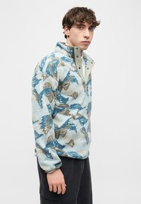 Νεαρός άνδρας που φοράει μπλε και μπεζ φλις πουλόβερ με μοτίβο, με κολάρο ανοιχτού φερμουάρ και σκούρο παντελόνι, στέκεται πλαγίως μπροστά σε απλό φόντο.