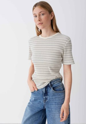 Giovane donna con capelli lisci castano chiaro, indossa una maglietta a maniche corte a righe bianche e jeans blu, in piedi con una mano in tasca.