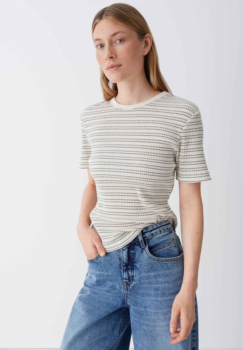 Jonge vrouw met recht lichtbruin haar, gekleed in een wit gestreept T-shirt met korte mouwen en een blauwe spijkerbroek, staand met één hand in de zak.