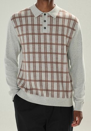Man wearing light gray long-sleeve polo sweater with brown plaid pattern and black buttons, paired with black pants.