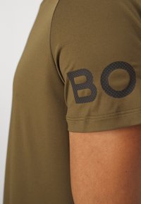 Krátké tričko olivově zelené barvy z hladké, prodyšné látky. Černé logo "BO" s tečkovanými akcenty na levém rukávu.