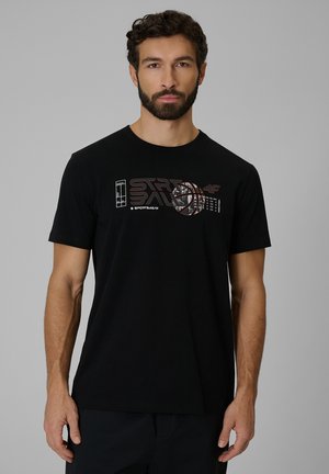 Mężczyzna z kręconymi włosami i brodą, ubrany w czarny t-shirt z grafiką koszykówki i napisem "Street Ball" na przodzie, na szarym tle.