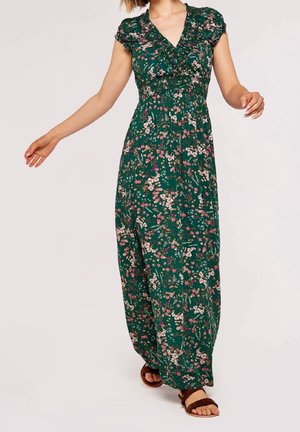 Femme portant une robe maxi verte à fleurs avec des manches courtes et un col en V, associée à des sandales plates marron, marchant avec les bras légèrement levés.